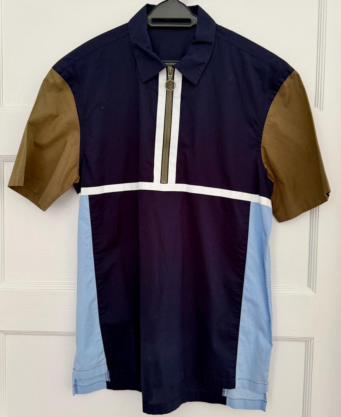 Navy blue polo shirt, khaki sleeves, light blue panels, and a zipper with white stripes. Hanging on a black hanger over a white door.