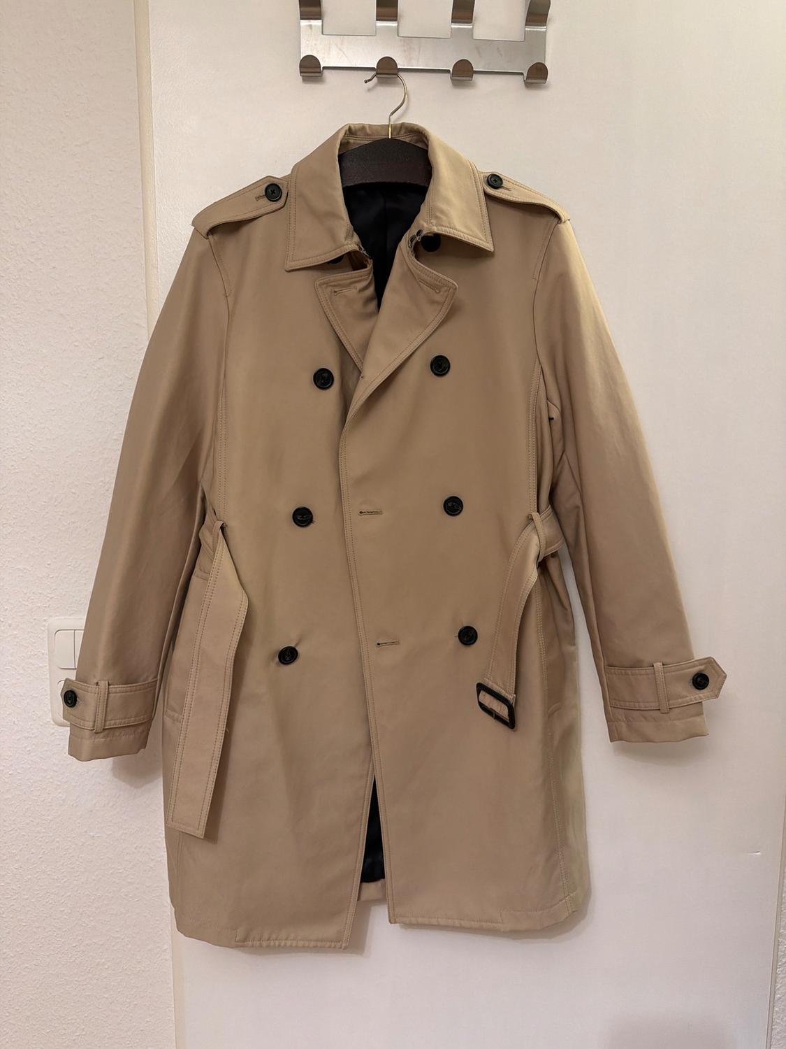 Beige trench coat hanging on a white wall hook, with black buttons and a belt. Minimalist and modern setting.