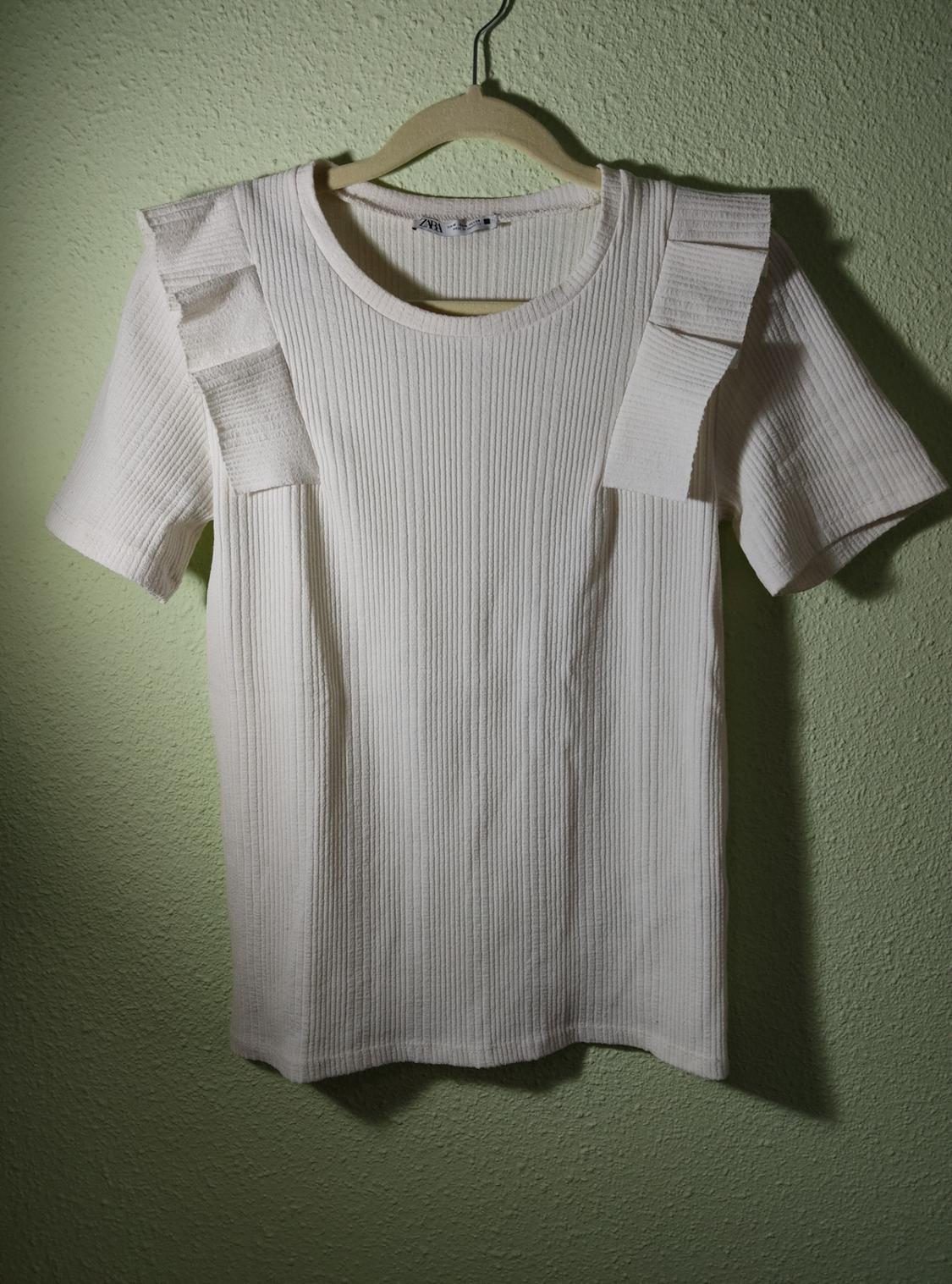 White ribbed T-shirt with frilled shoulders, hanging on a hanger against a light green wall with shadows. ZARA label.