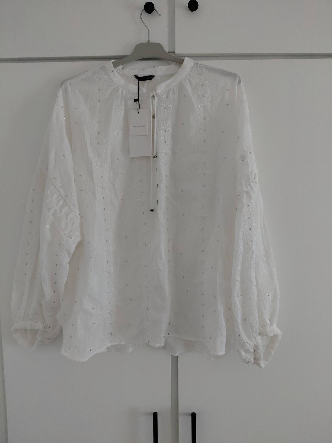 White blouse with embroidery, shiny details and a bow at the neck, hanging on a hanger in front of a white wardrobe.