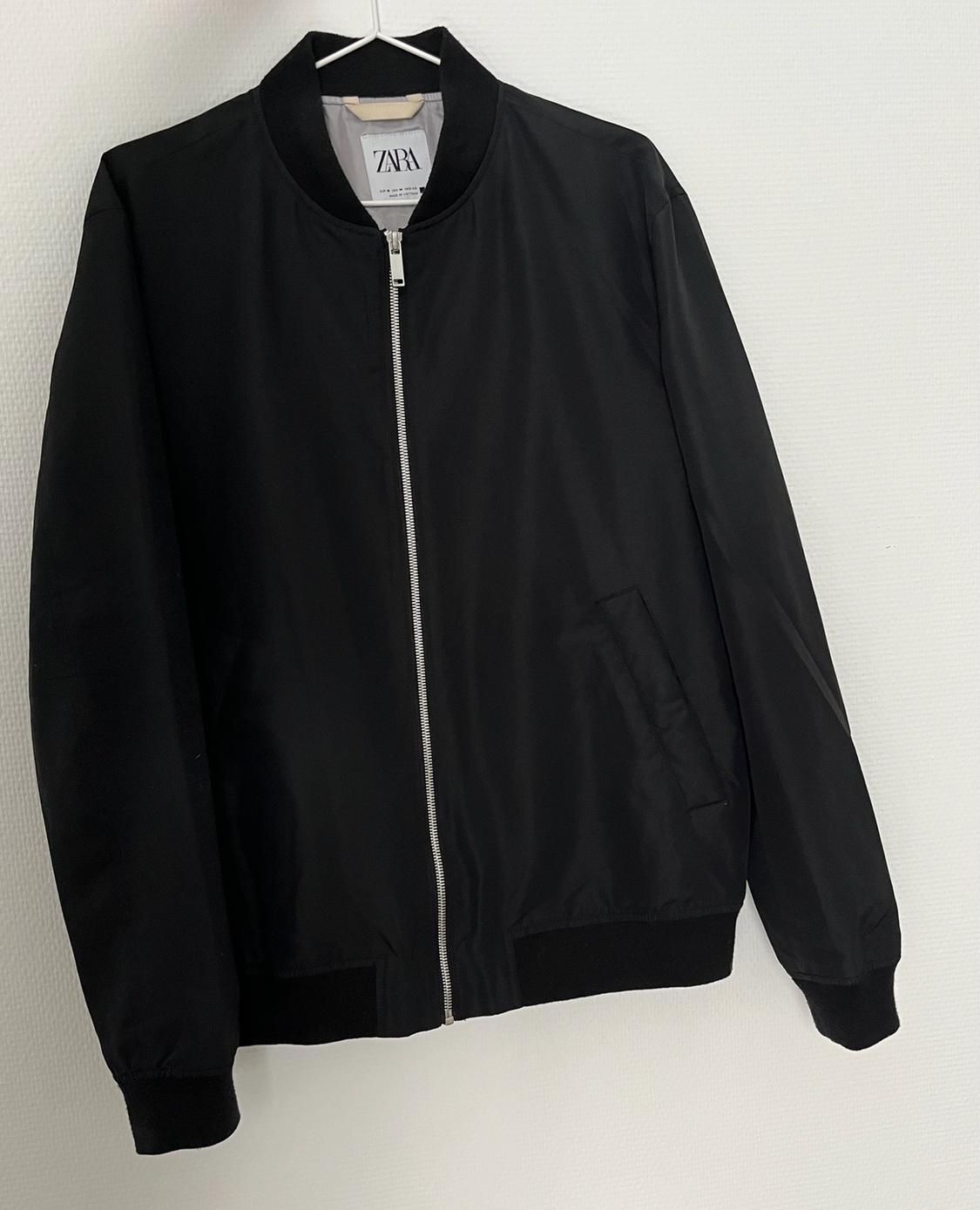 Black bomber jacket with a front zip hanging on a hanger against a white wall. Inner label with the text ZARA.