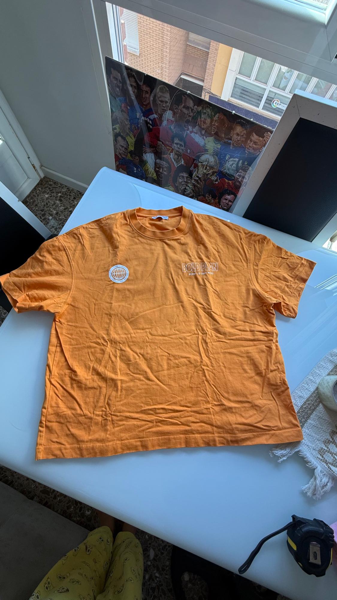 Orange T-shirt on a white table with a patch and the text BOLD PLAN READY TO TAKE RISKS. Casual indoor setting with a window and a poster.