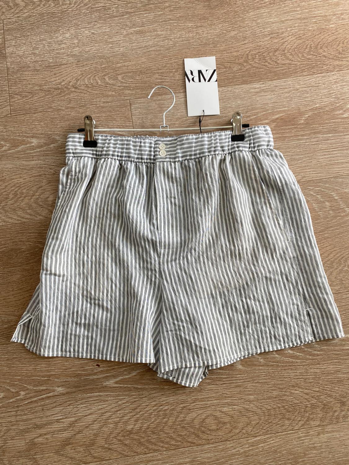 Gray and white striped shorts hanging on a hanger over a wooden floor. Visible tag with text "ZARA".