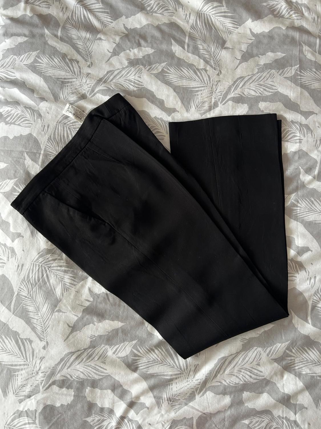 Black trousers on a bed with a grey and white leaf-patterned duvet. Visible label on the waistband of the trousers.