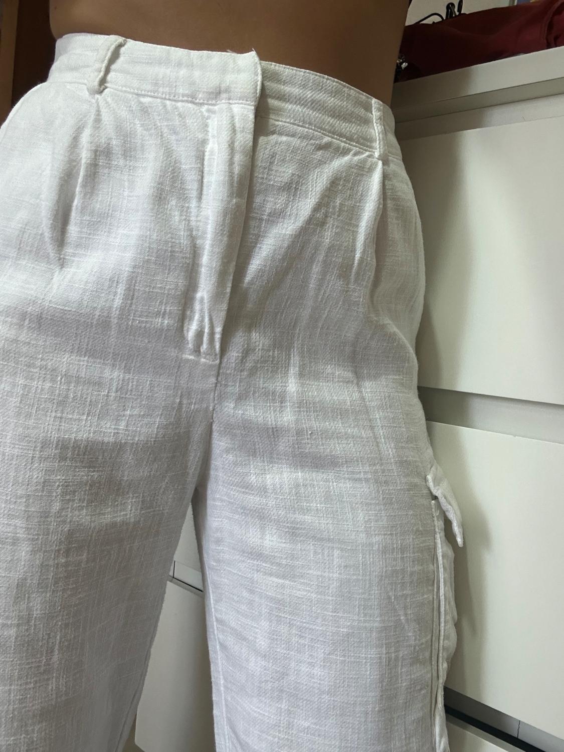 White linen trousers with pleats, seen close up. Background with white chest of drawers and red handbag.