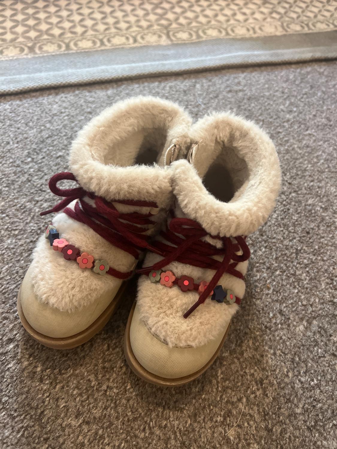Children’s beige boots with a faux-fur texture, burgundy laces and flower beads, photographed on a grey rug.