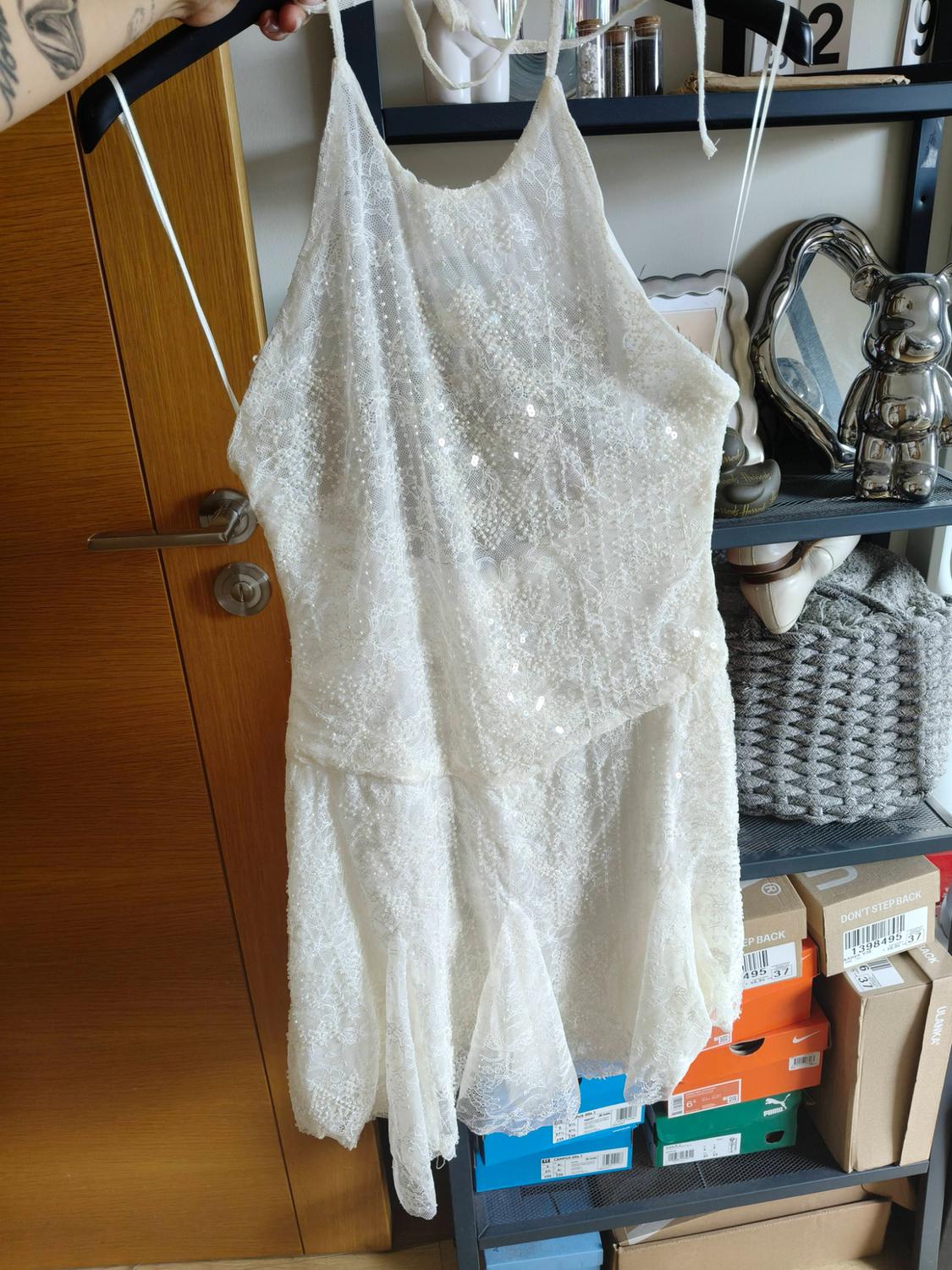 Hand holding a white lace dress on a hanger. In the background, shelving with boxes and the text DON'T STEP BACK, ULANKA.