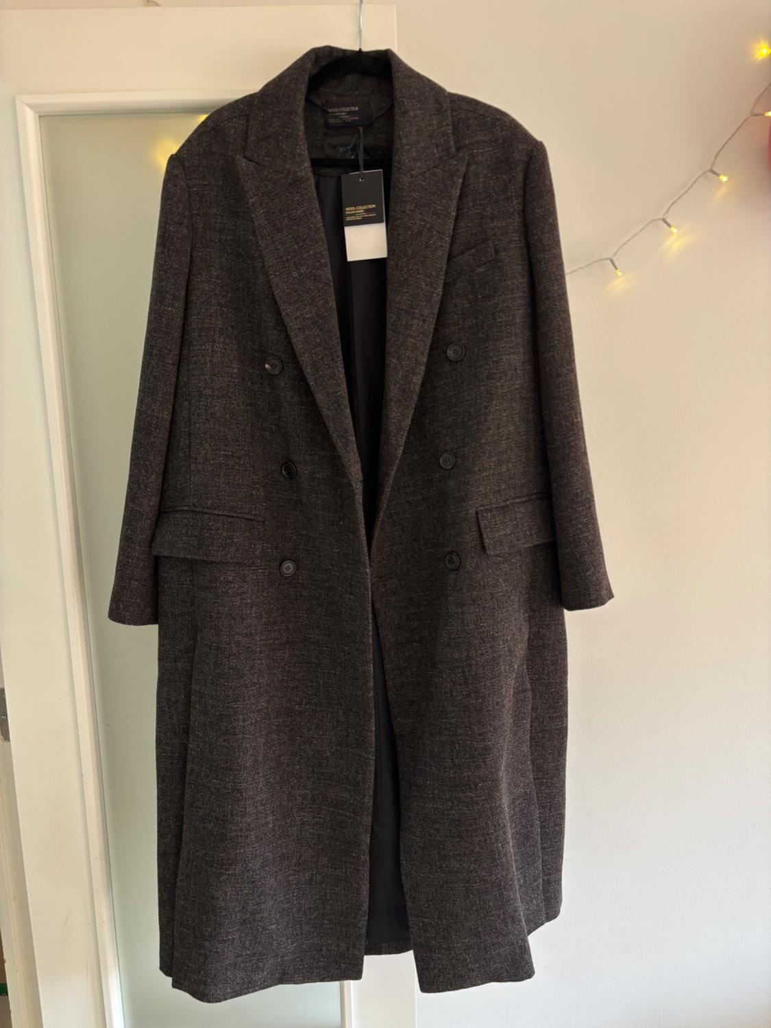 Dark grey long coat hanging on a white door, with decorative lights in the background. Label visible on the collar.