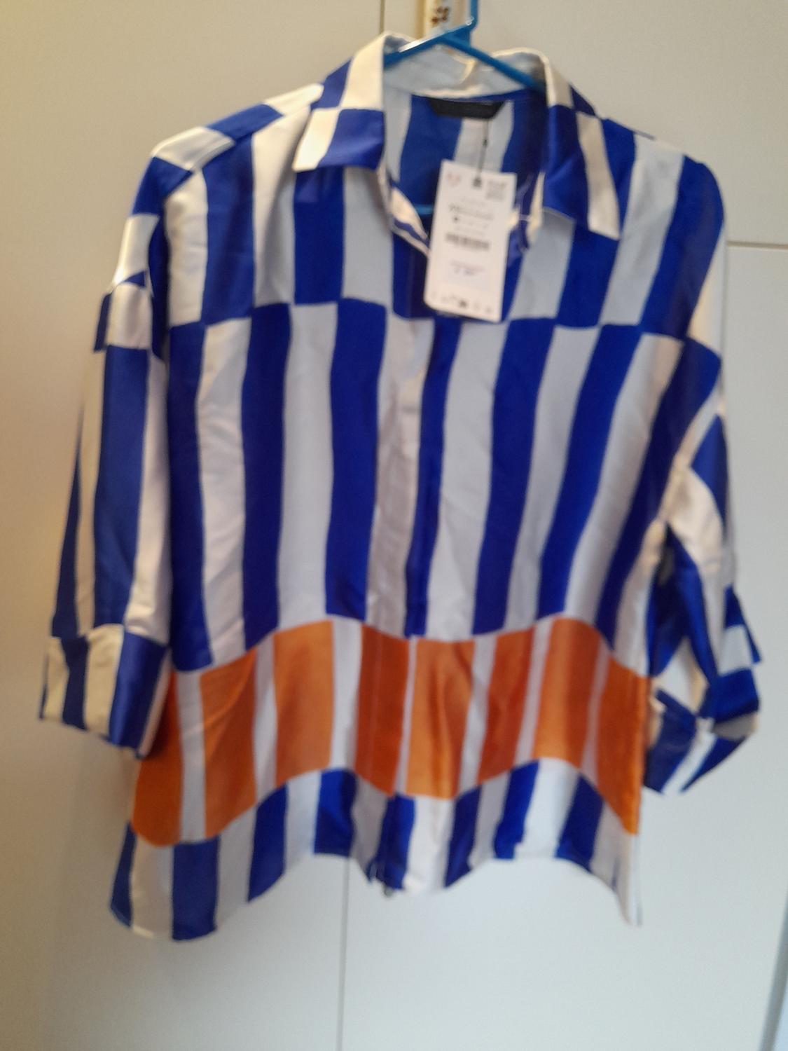 Μauρόbluza μακρυμάνικη με μπλε και λευκές κατακόρυφες ρίγες και μια πορτοκαλί λωρίδα στο κάτω μέρος, κρεμασμένη σε κρεμάστρα.
