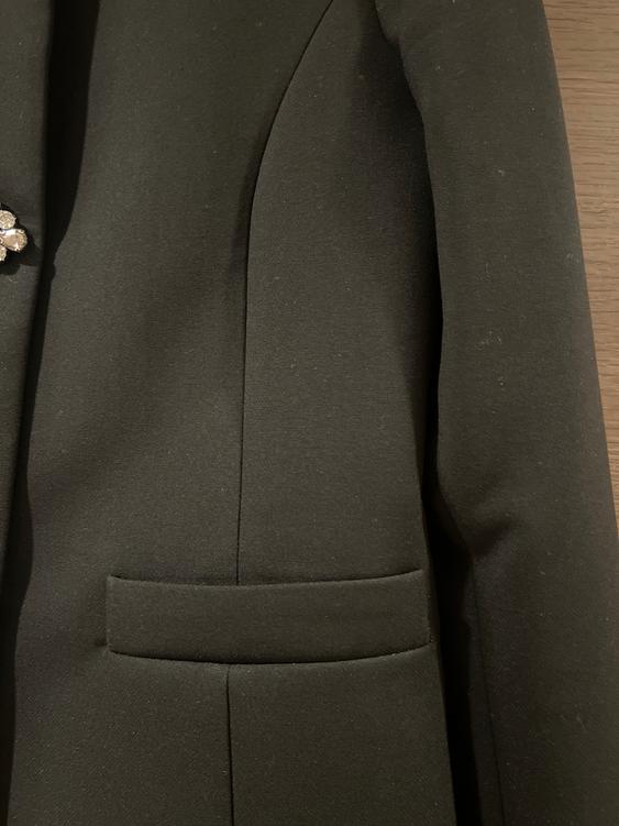Detail of a black jacket with a lapel and pocket, on a wooden background. A decorative brooch is visible on the left side.