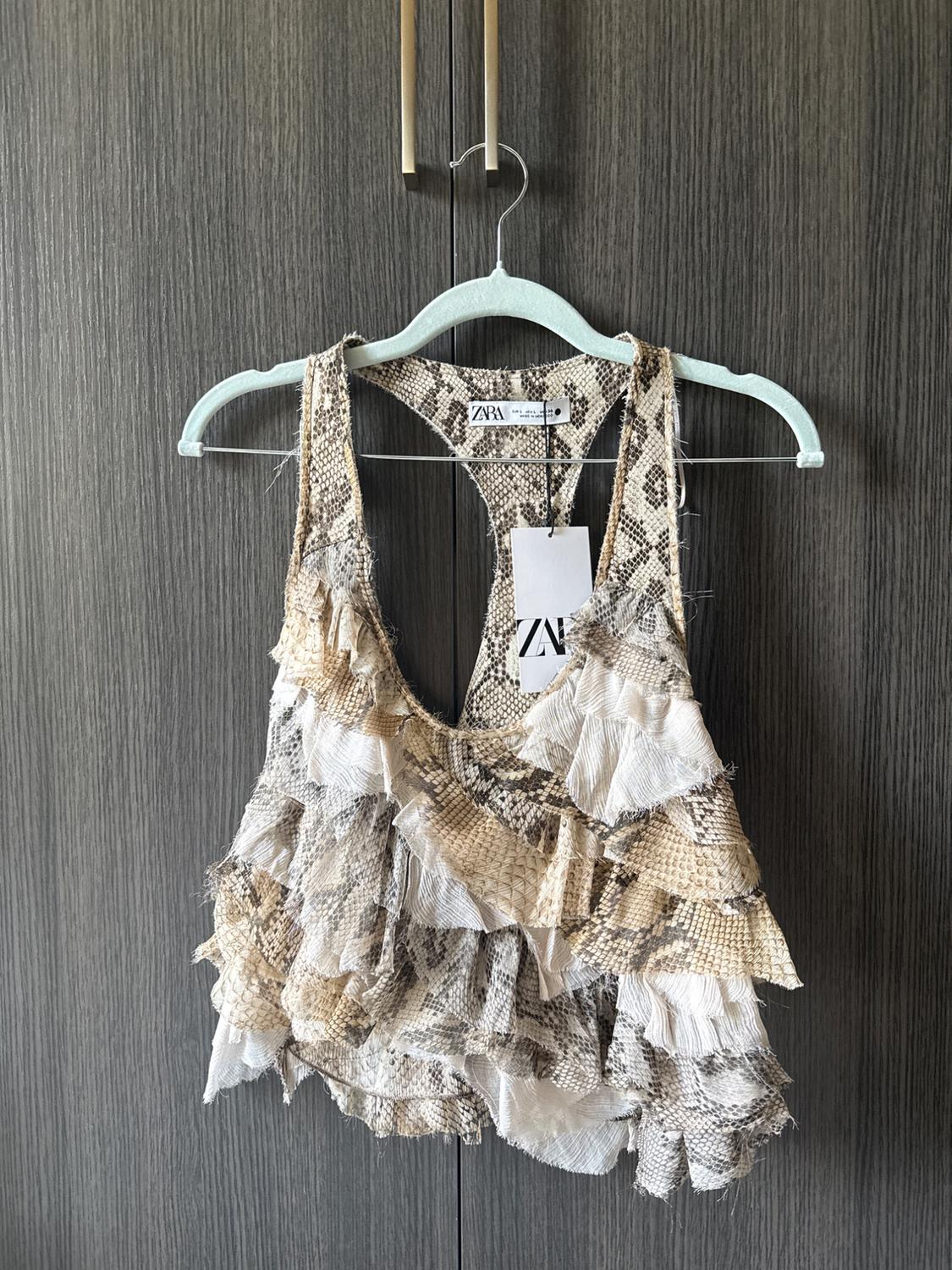 Snake-print strappy top with frayed ruffles hanging on a hanger in front of a wooden wardrobe. Text: ZARA.