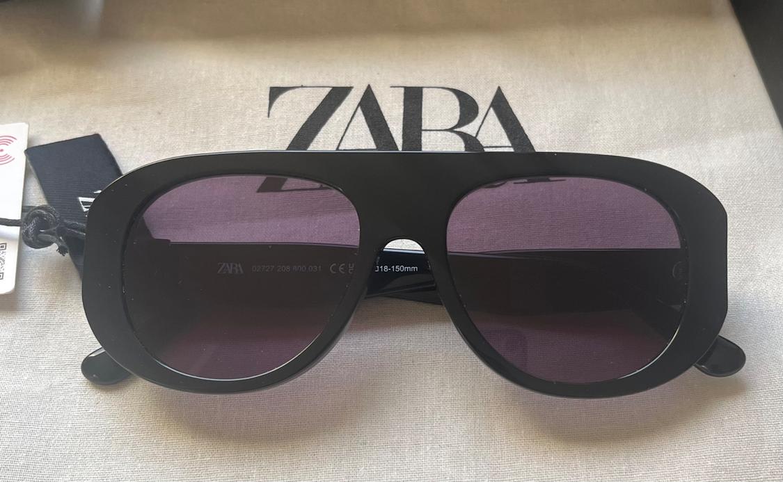 Black sunglasses on fabric. Text: ZARA, ZARA 02727 208 800 031 CE 18-150mm.