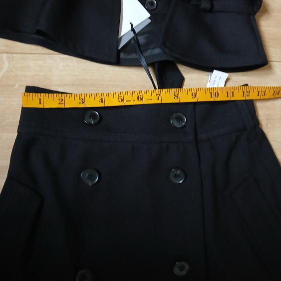 Black skirt with double buttons and a yellow tape measure around the waist. Light wood background. Visible tags.