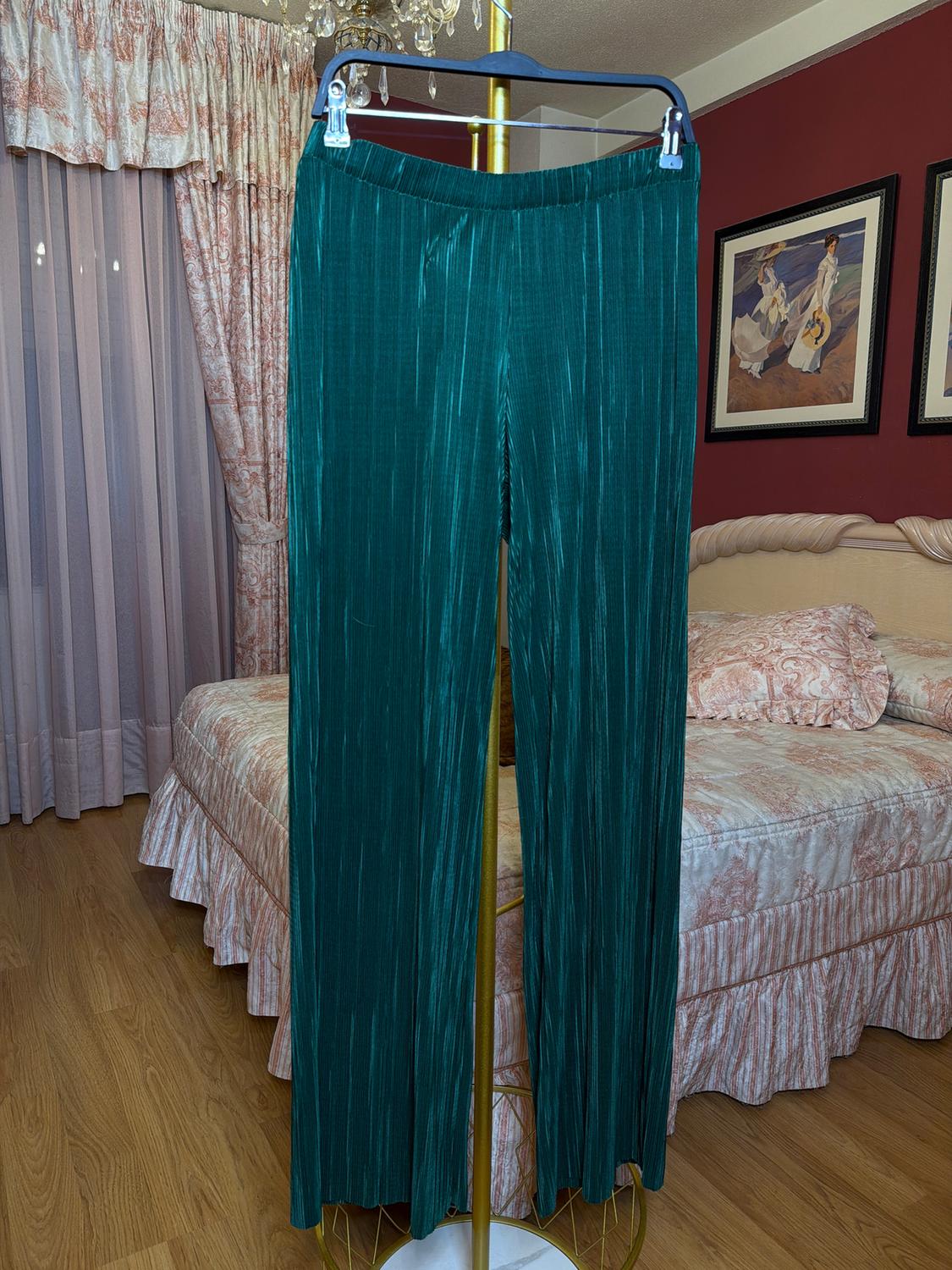 Green pleated trousers hanging on a hanger in a bedroom with curtains and a bed with a patterned bedspread. Classic decor.