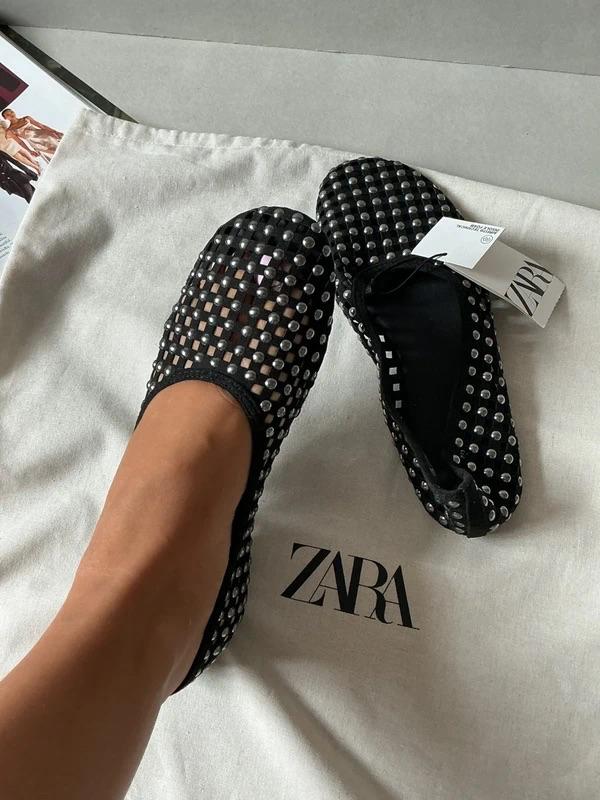 Foot wearing a black mesh flat shoe with silver studs next to its pair on a fabric bag. Visible text: ZARA.