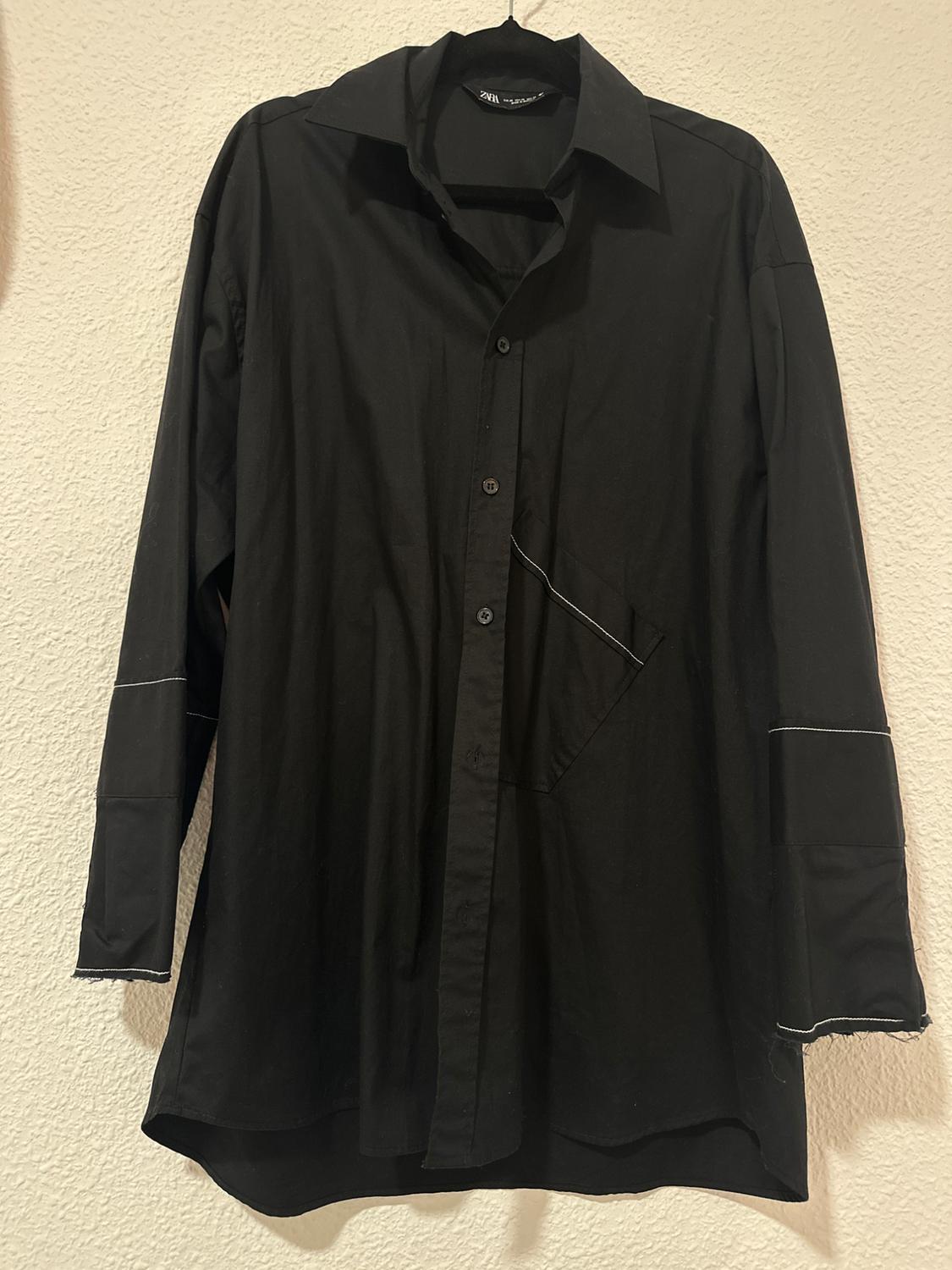 Black long-sleeved shirt hanging on a hanger, with white stitching details on the sleeves and a side pocket.