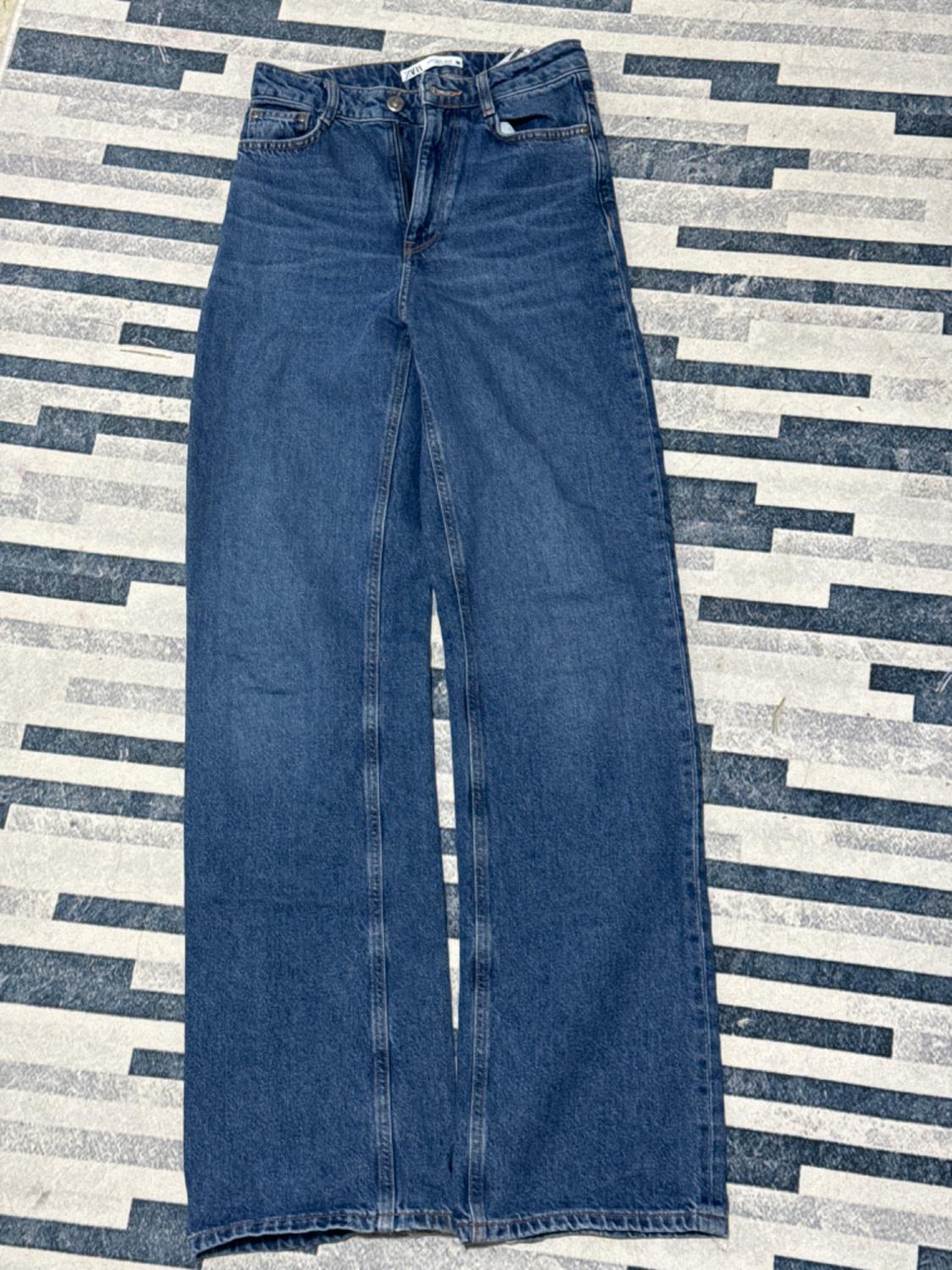 Wide-leg μπλε τζιν πάνω σε ένα χαλί με γεωμετρικό σχέδιο σε αποχρώσεις του γκρι και του μπλε.