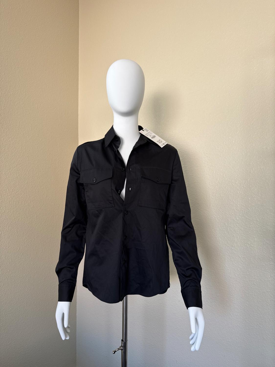 Mannequin wearing a black long-sleeve shirt with front pockets, in a minimalist setting with light walls. Visible tag on the collar.