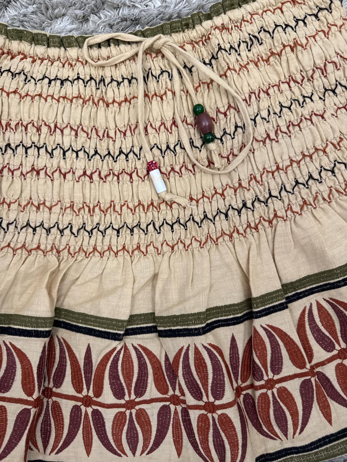 Detail of a beige skirt with a ruched waist, beaded drawstring, and leaf design in reddish-brown tones on a grey rug.