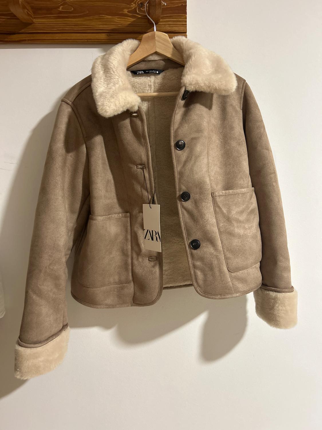 Beige jacket with a wool collar and cuffs, hanging on a wooden coat hanger. The visible label displays the text "ZARA".