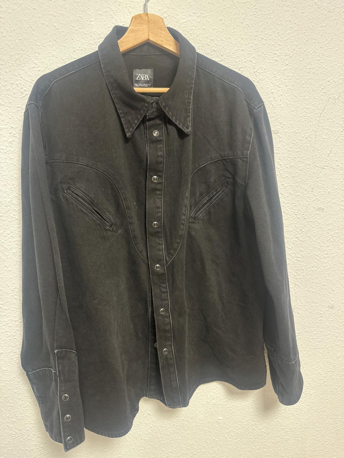 Black denim shirt hanging on a wooden hanger, with metallic buttons and a visible Zara label on the collar.