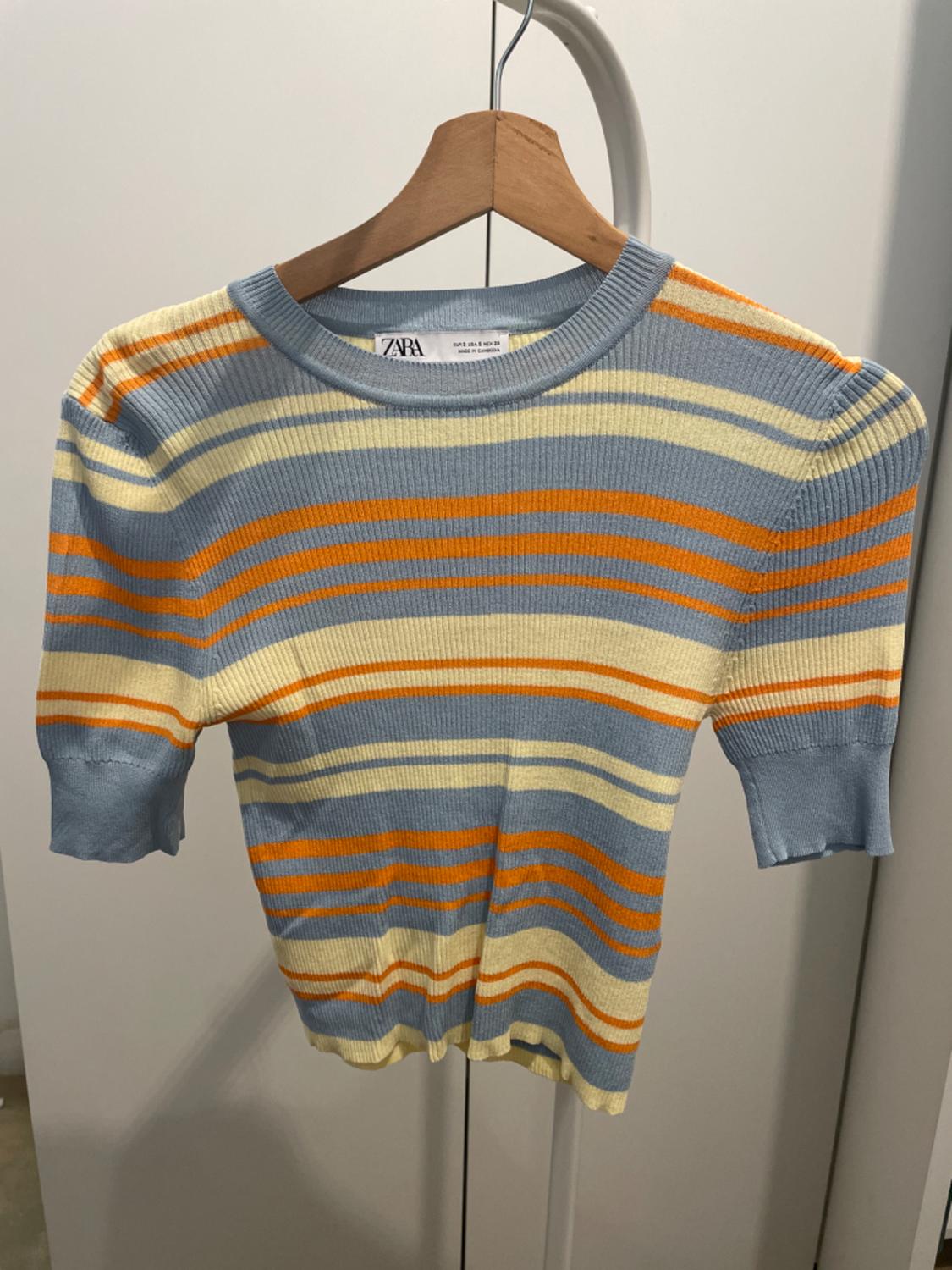 Blue, orange, and cream striped jumper hanging on a wooden hanger. Visible label with the text "ZARA". White wardrobe background.