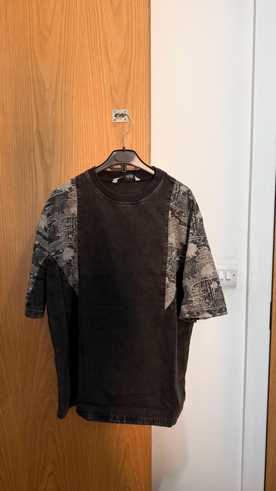 Casual black t-shirt with patterned panels, hanging on a hanger over a wooden door next to a white wall.