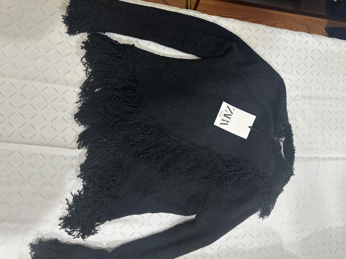 Black fringed jacket on a white surface with a diamond pattern. Visible label with text: "ZARA".