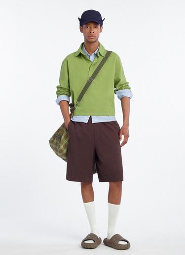 Modelo con gorra azul, suéter verde, camisa a cuadros, bermudas marrones, calcetines blancos y sandalias. Lleva bolso de cuadros.