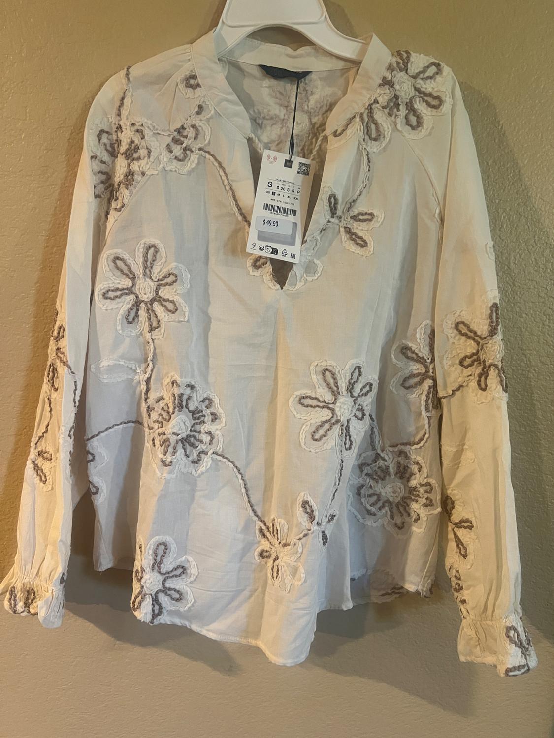 White blouse with floral embroidery in shades of brown and beige, hanging on a hanger. Visible tag with price of $49.90.