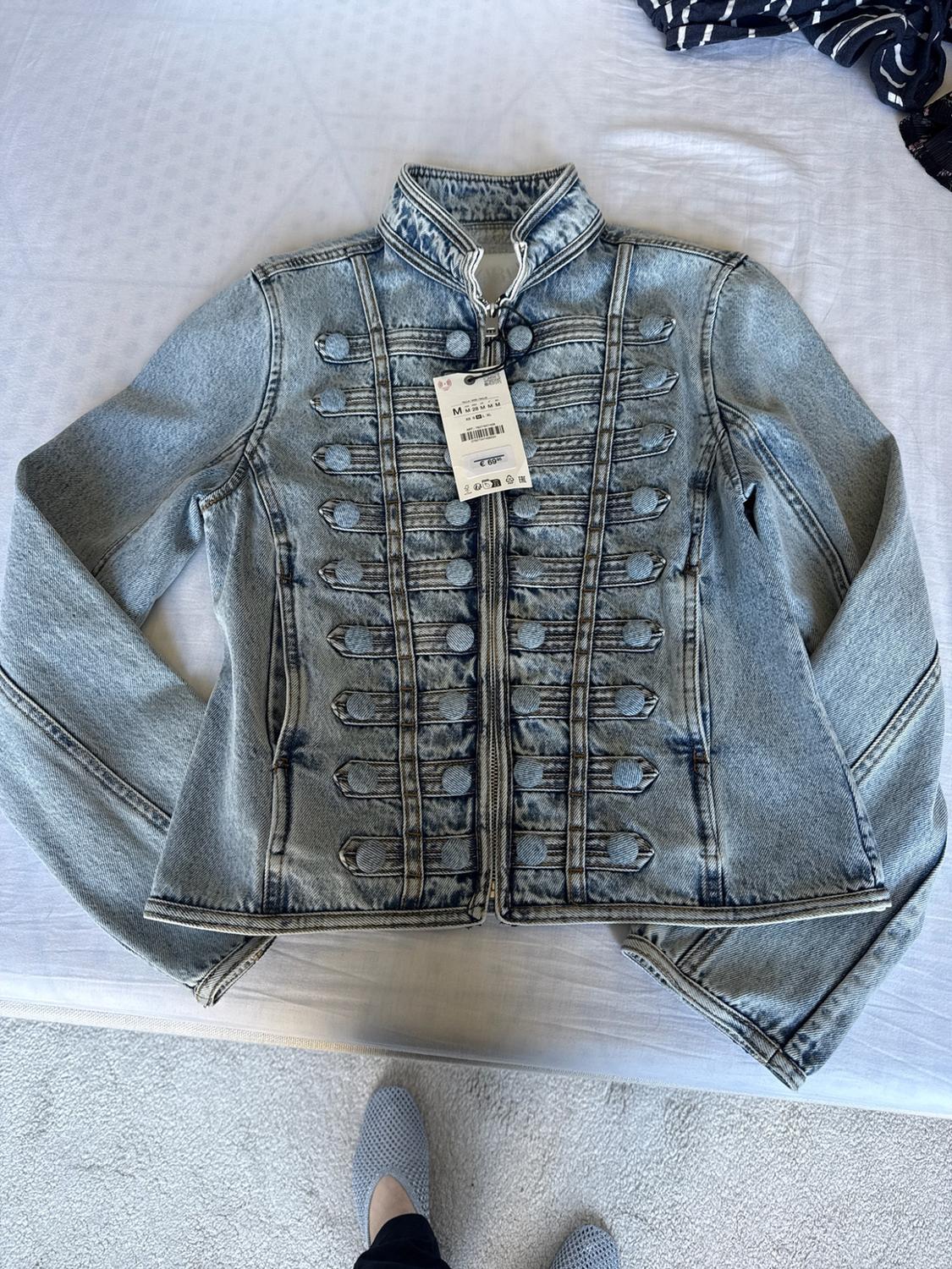 Blue denim jacket, military style, with buttons, zip and label, on a white bed. Below, a foot in a grey shoe is visible.