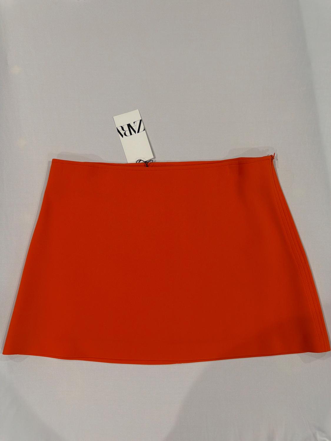 Top-down view of a plain orange miniskirt on a white background. There is a label on the waistband with the text ZARA.