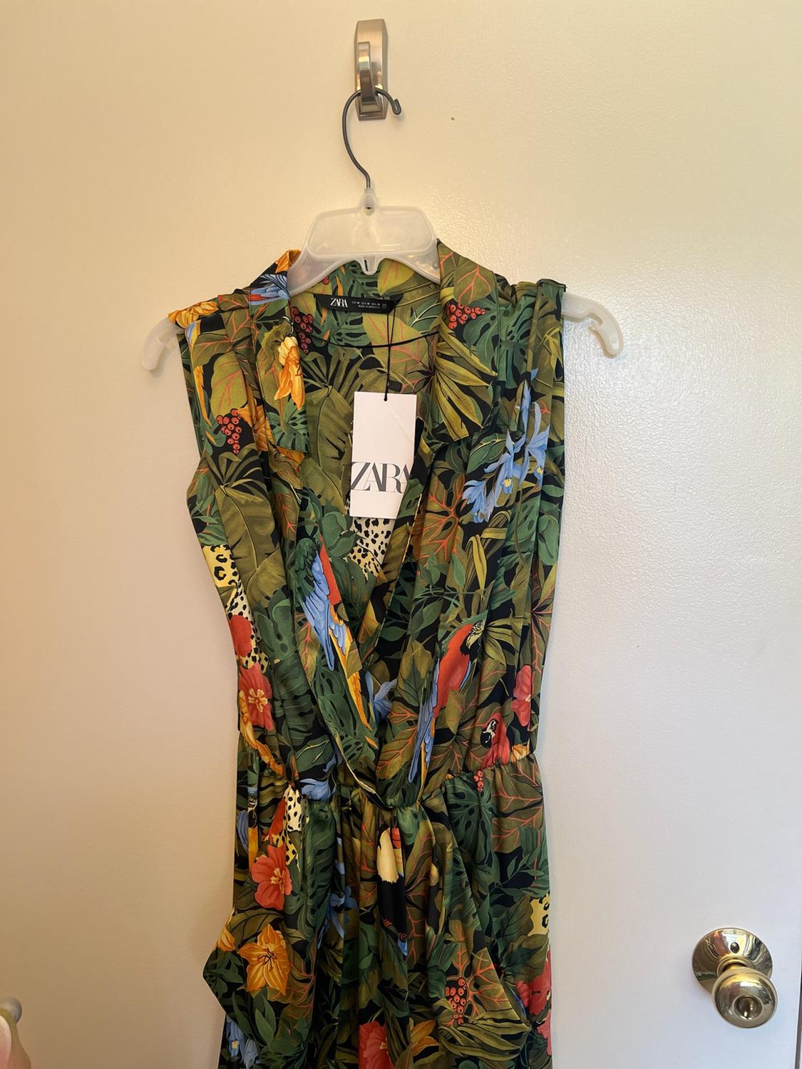 Sleeveless dress with a tropical leaf and bird print, hanging on a white door. Visible label with the text ZARA.