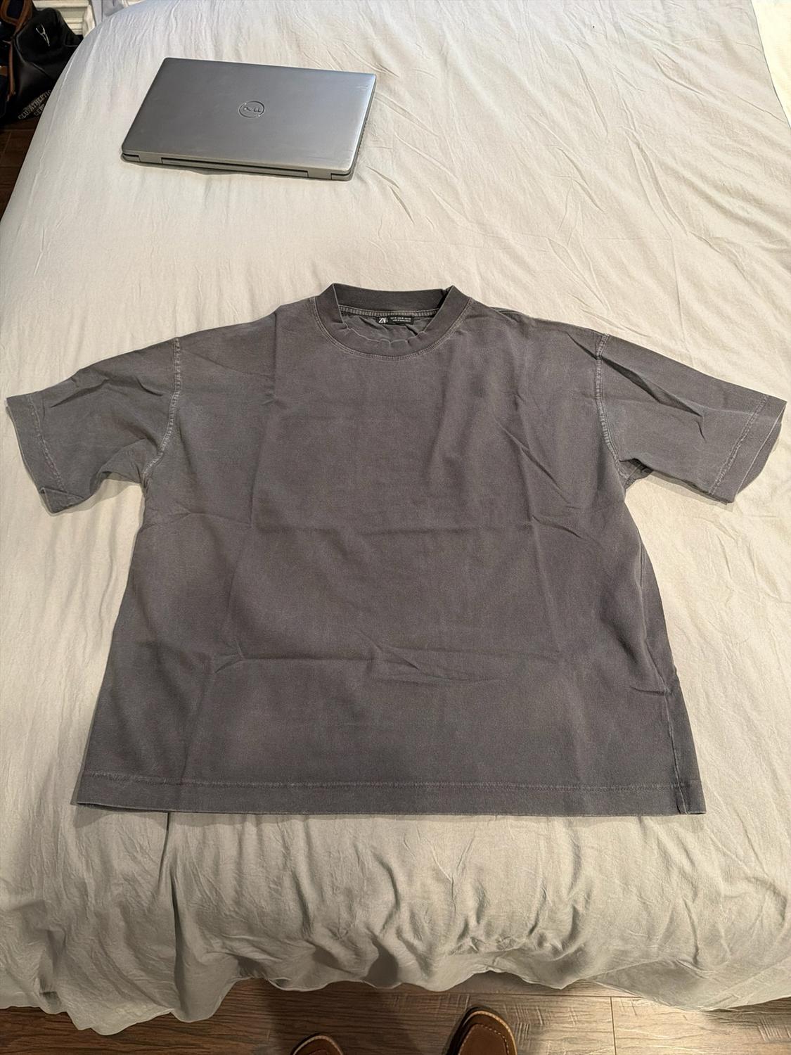Dark grey short-sleeved T-shirt on a bed with light-coloured sheets. In the background, a closed laptop. Casual and relaxed style.