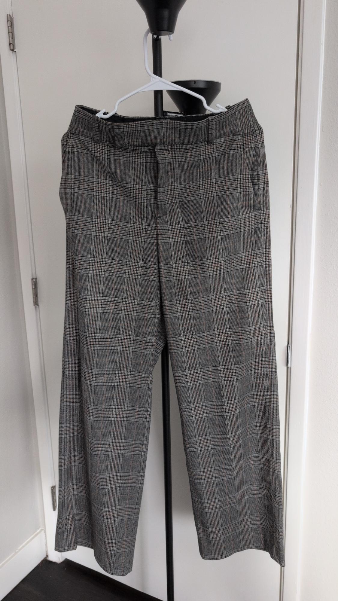 Gray plaid pants hanging on a white hanger on a black stand in front of a white door in an indoor setting.