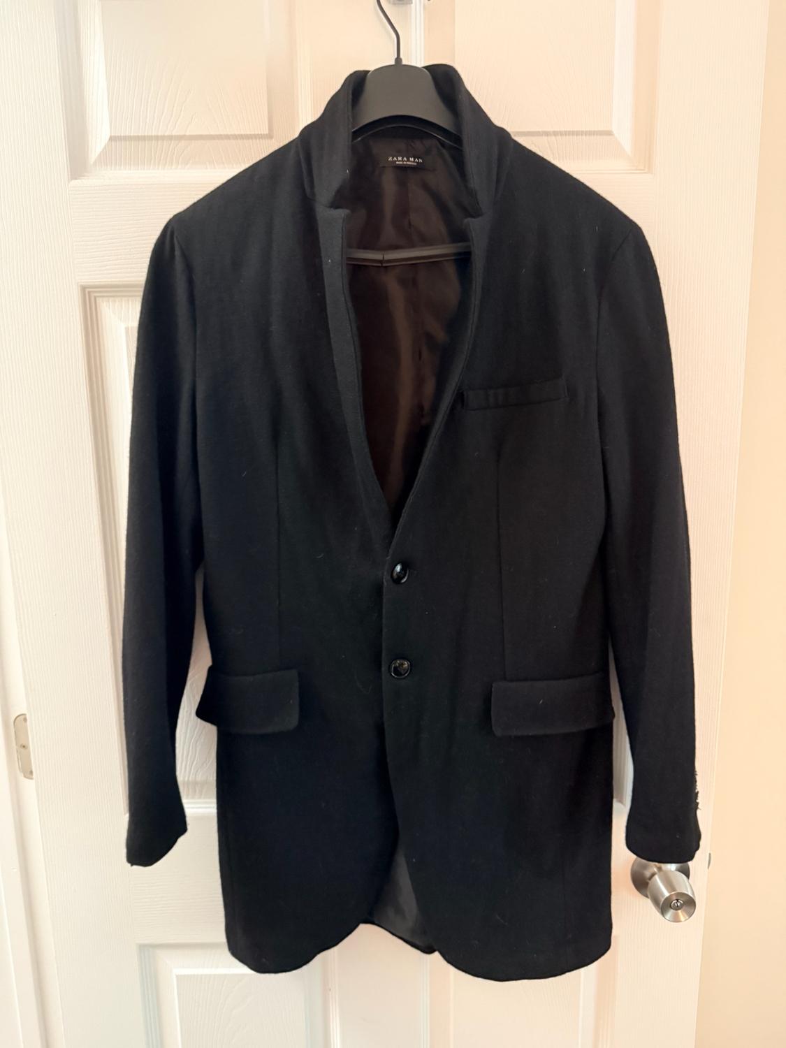 Black two-button blazer with flap pockets, hanging on a white door. The inner label reads ZARA MAN.