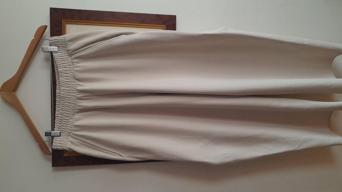 Beige trousers hung on a wooden hanger, with an elasticated waist, against a white wall background and picture frame.