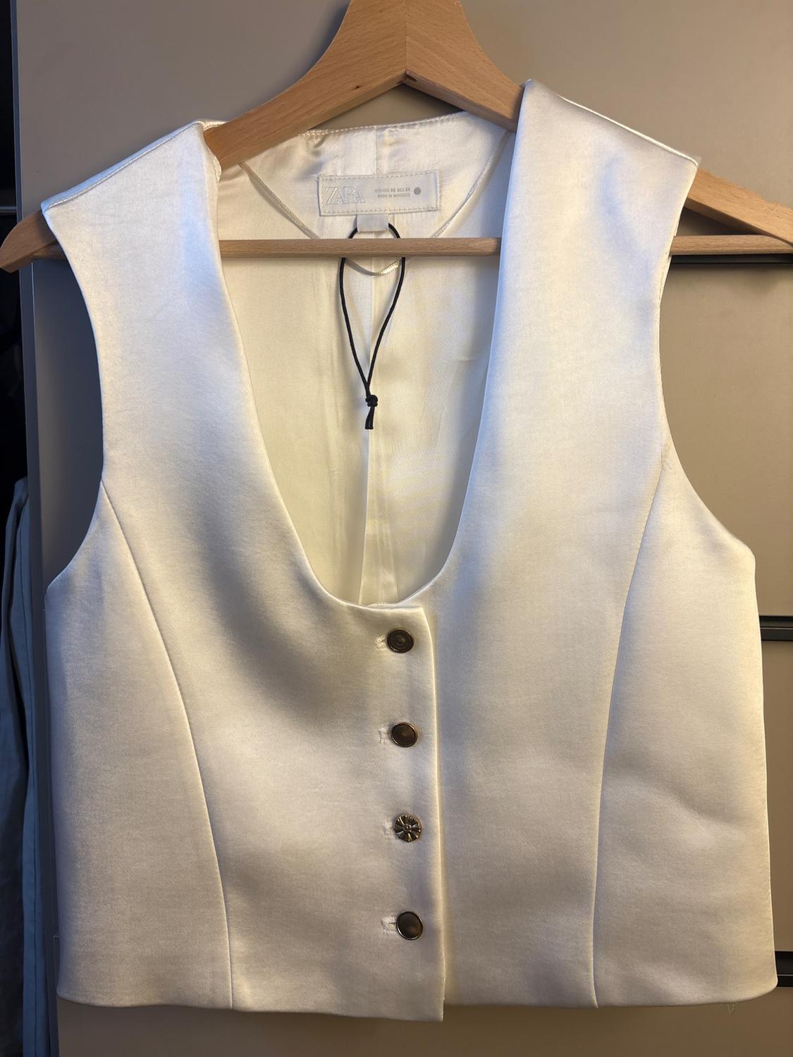 White sleeveless waistcoat with brown buttons, hanging on a wooden hanger. Label visible on the inside.