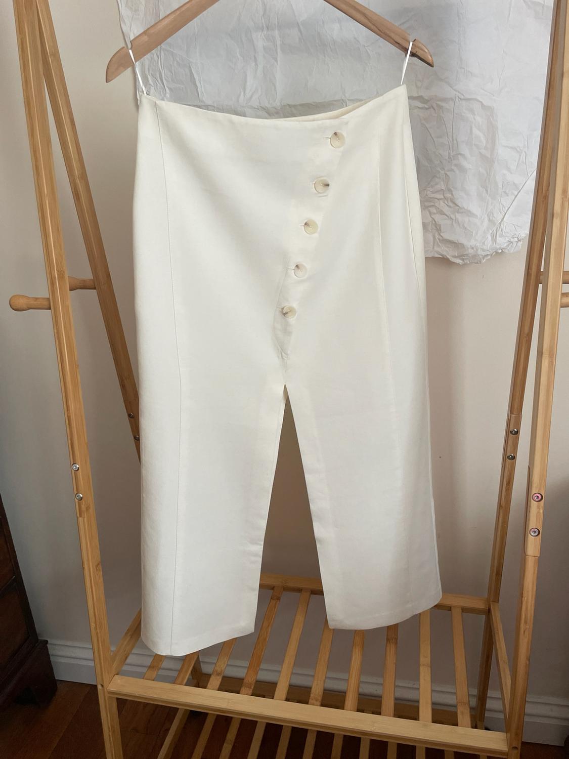White skirt with asymmetric buttons and a front slit, hanging on a wooden clothes rack against a light background.