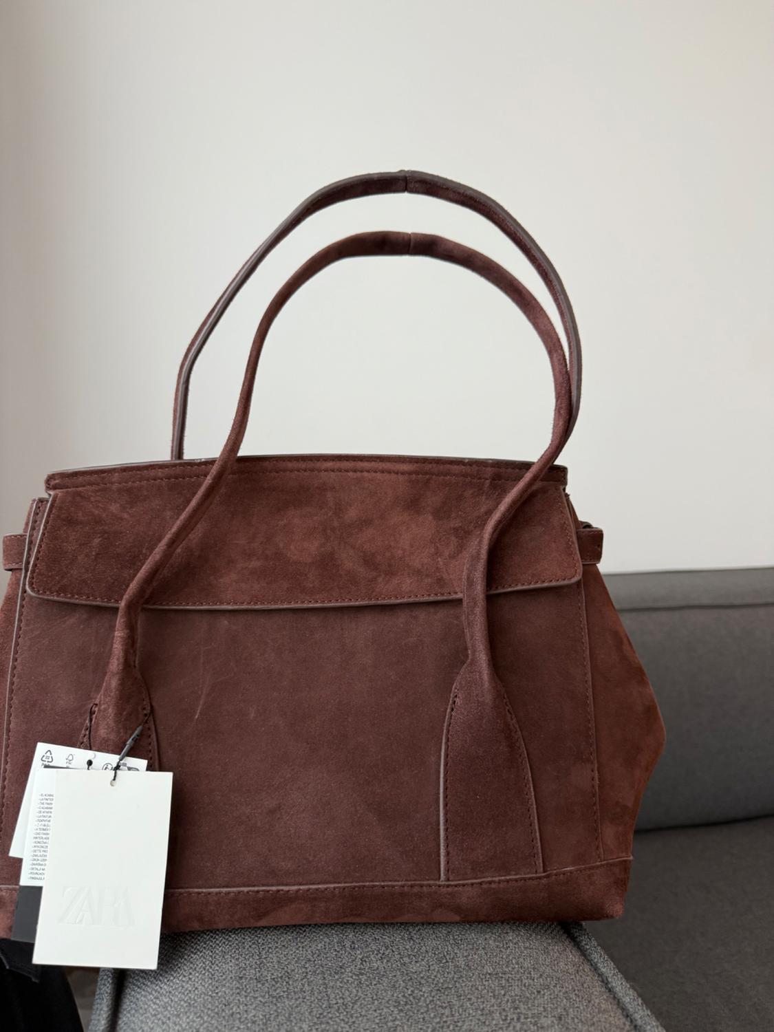 Brown suede handbag with double handles on a grey sofa. White hanging tag with the text "ZARA". White background.