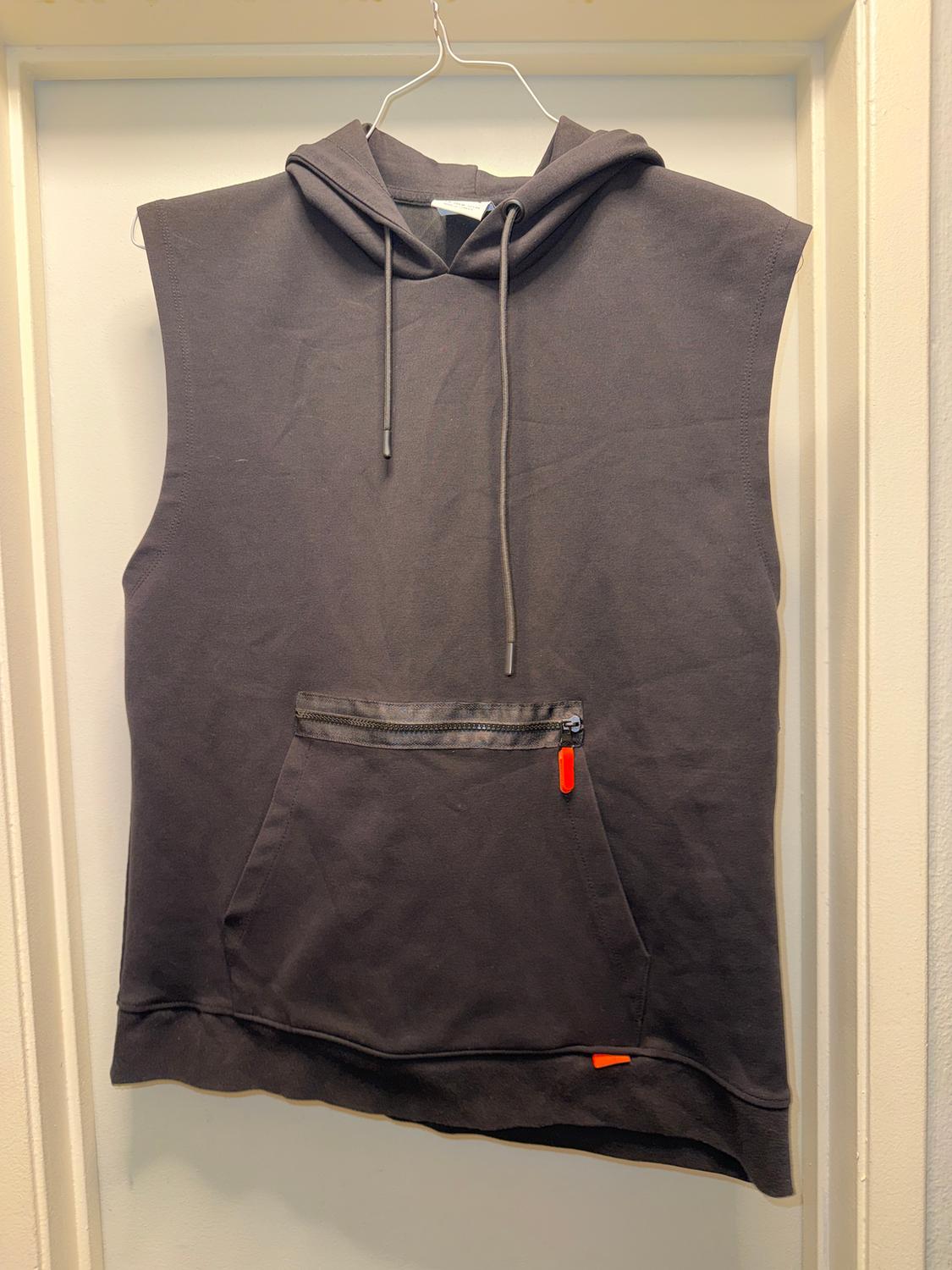 Black sleeveless hoodie with a front zipper pocket, hanging on a white hanger on a white door.