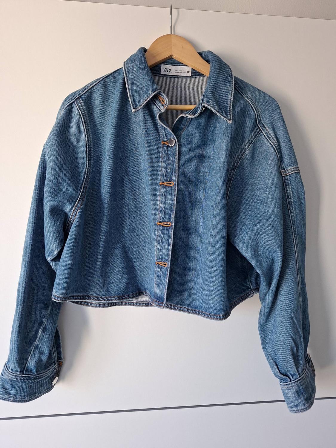 Short blue denim jacket with buttons, hanging on a wooden hanger in front of a white wall. ZARA label on the collar.