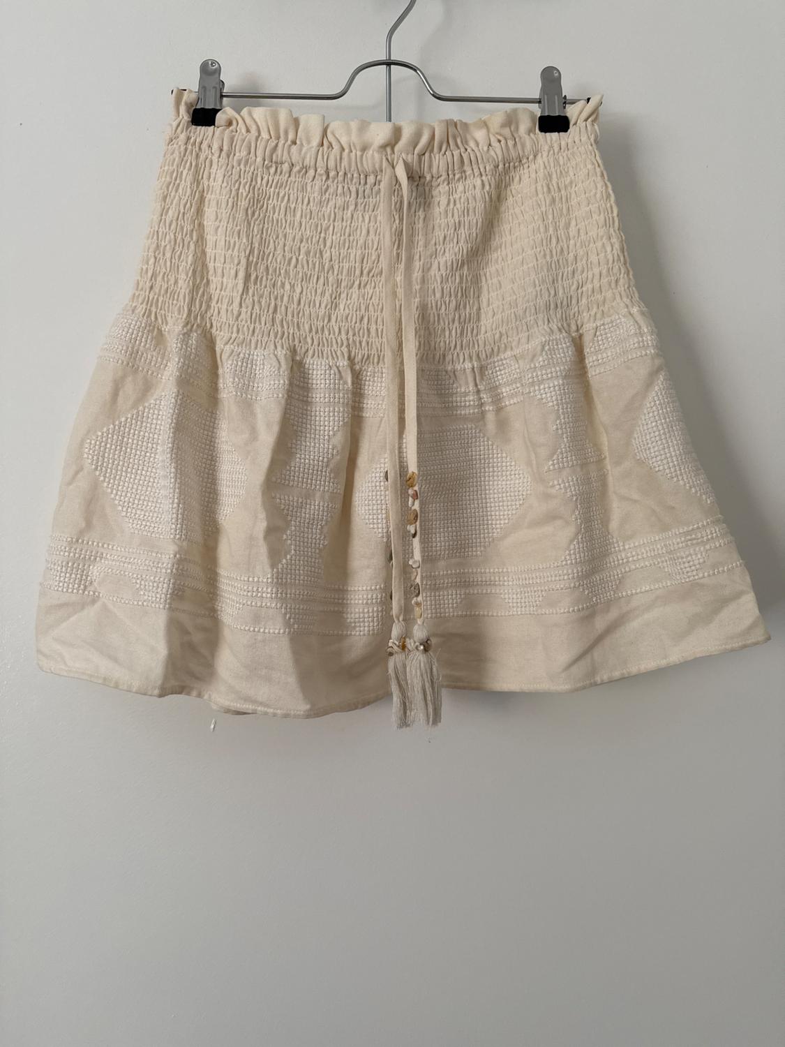 Cream-coloured short skirt on a hanger against a white background. Features a gathered elasticated waist, embroidery and a drawstring with shells and tassels.