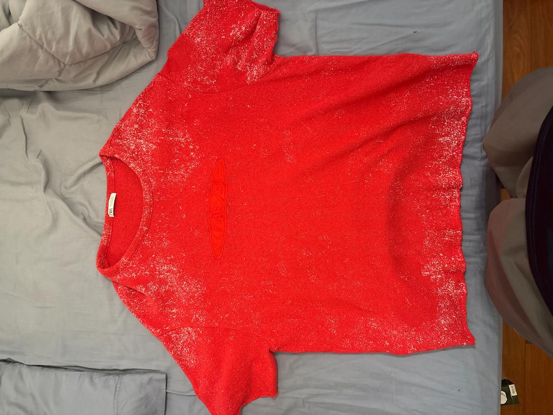 Red T-shirt with white mottled texture, placed on a bed with grey sheets. Label visible on the collar.