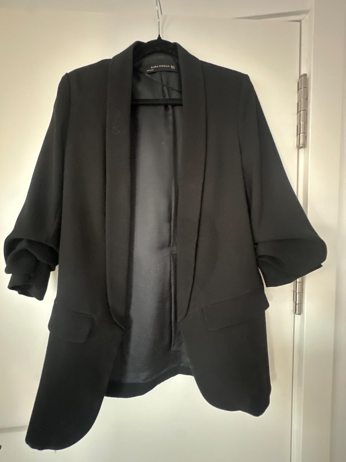 Black jacket hanging on a hanger over a white door. Rolled-up sleeves and wide lapels. Visible label on the inside.