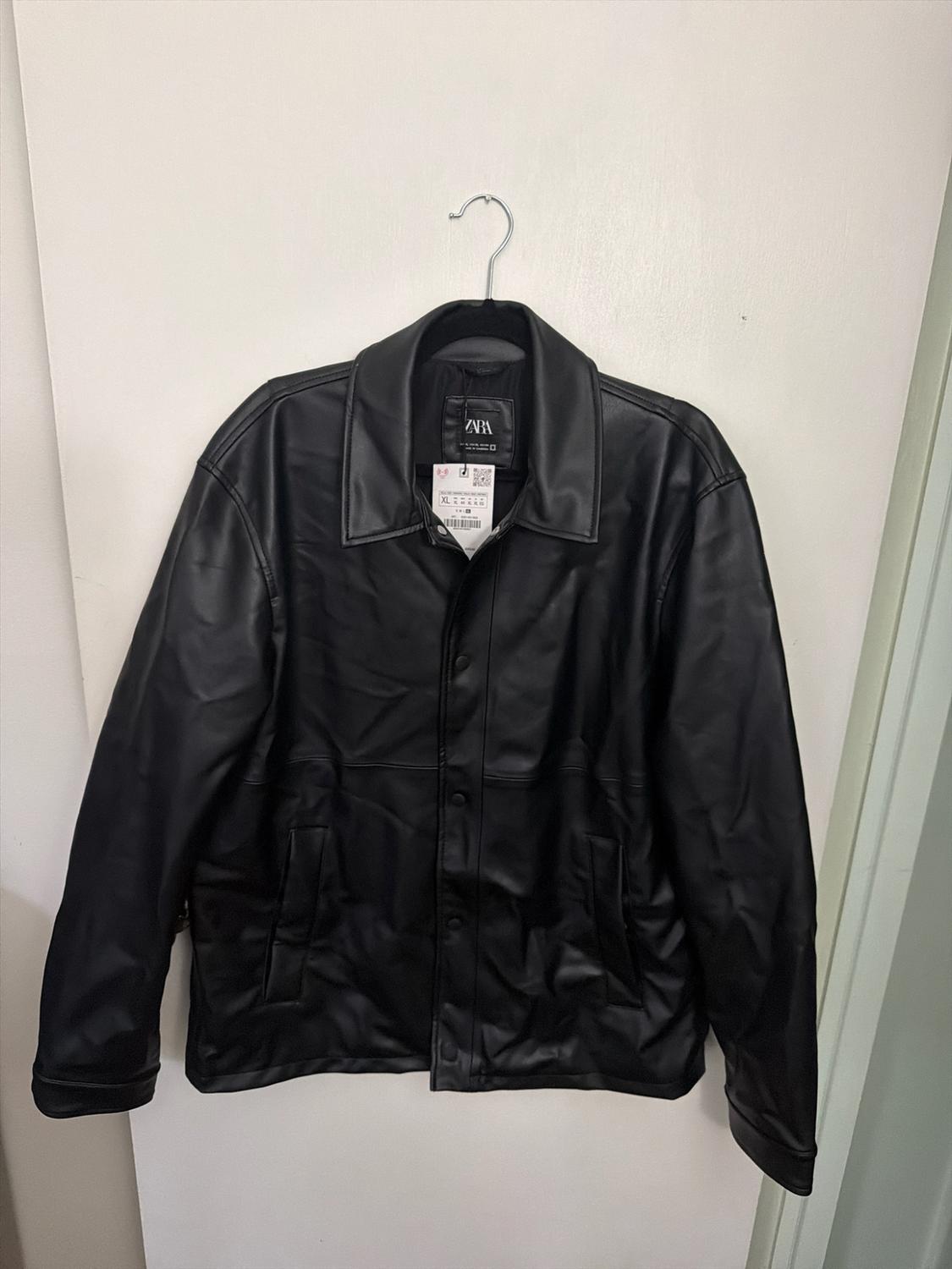 Black jacket hanging on a hanger over a white door. It has a collar, a button-front closure, and tags with the text ZARA and XL.