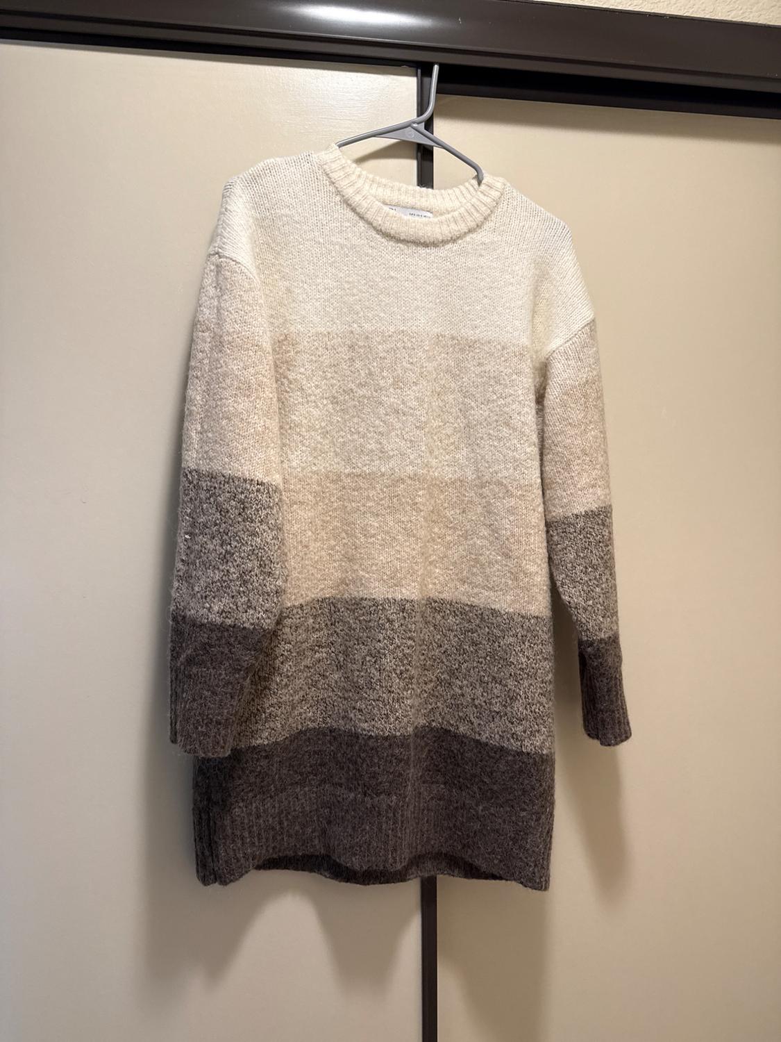 Long knit sweater with horizontal stripes in shades of white, beige, and brown, hanging on a hanger in front of a closet.