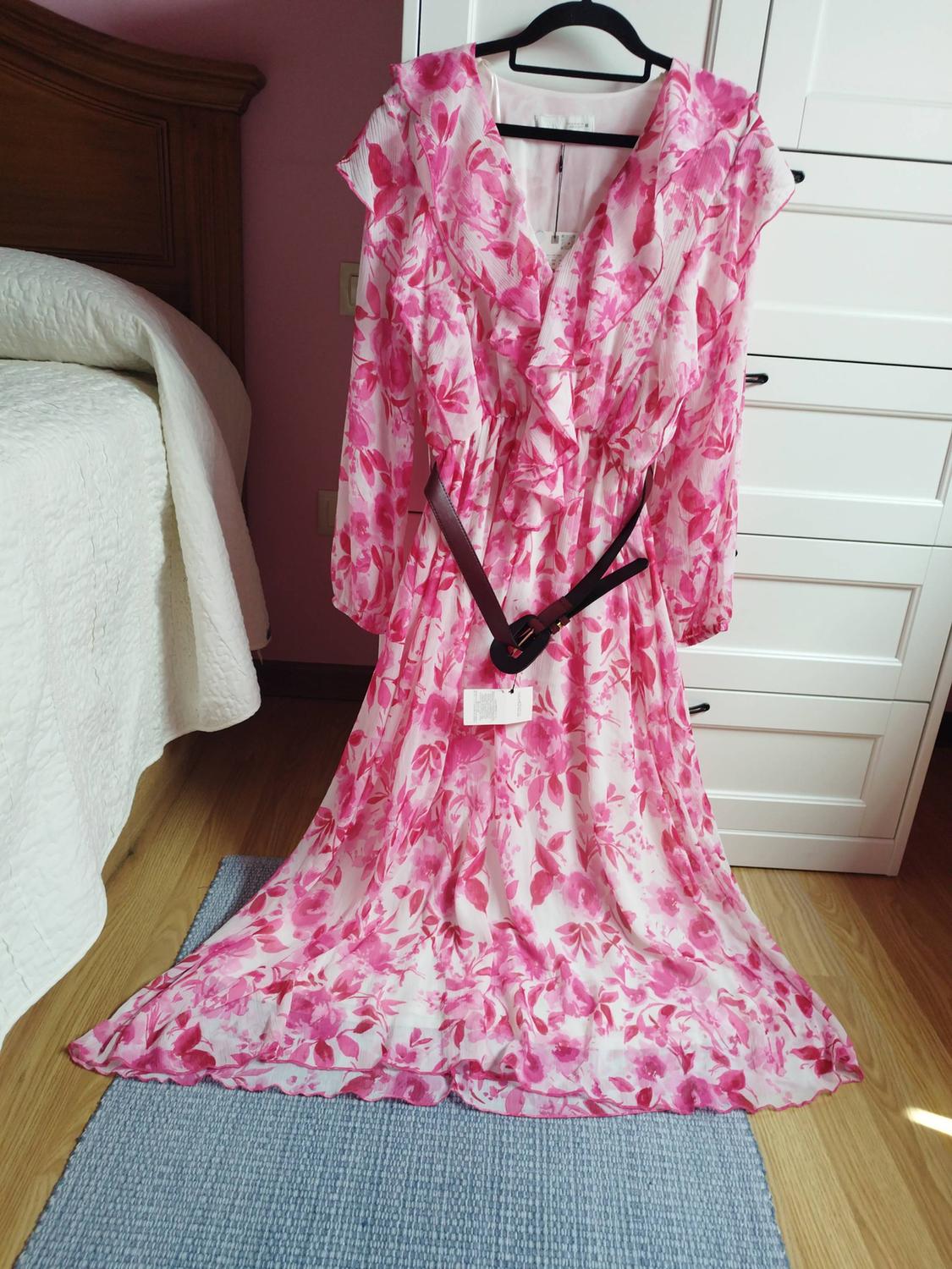 Long white dress with pink floral print, ruffles, and a dark belt, hanging on a hanger in a bedroom next to a bed.