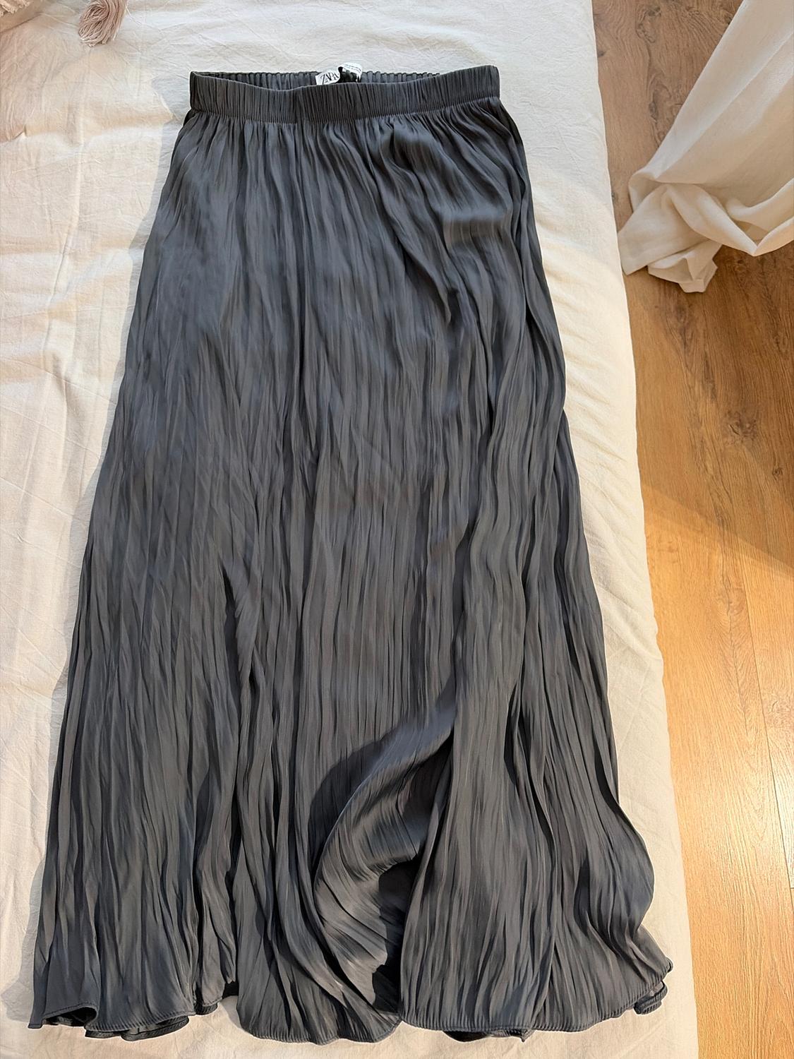 Dark grey pleated maxi skirt on a white bed, with wooden flooring visible in the background. Label visible on the waistband.