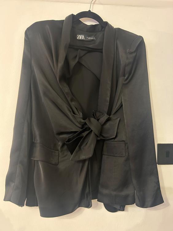 Satin black jacket with a large bow on the front, hanging on a hanger. Visible label with the text "ZARA".